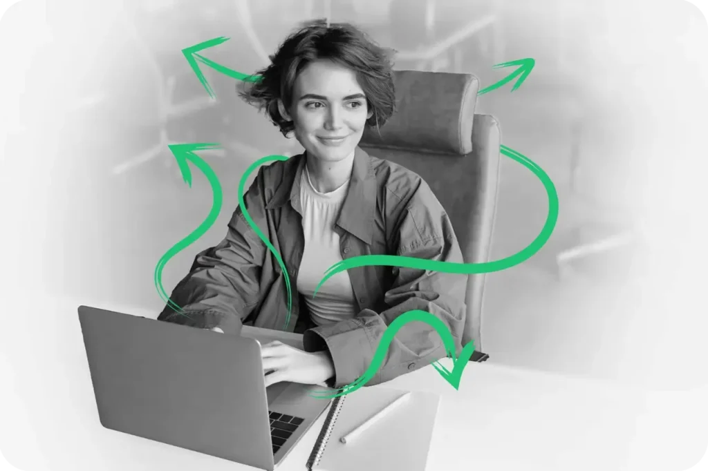 A black and white image of a smart casual dressed smiling young lady sitting at a desk with a laptop looking off to the side