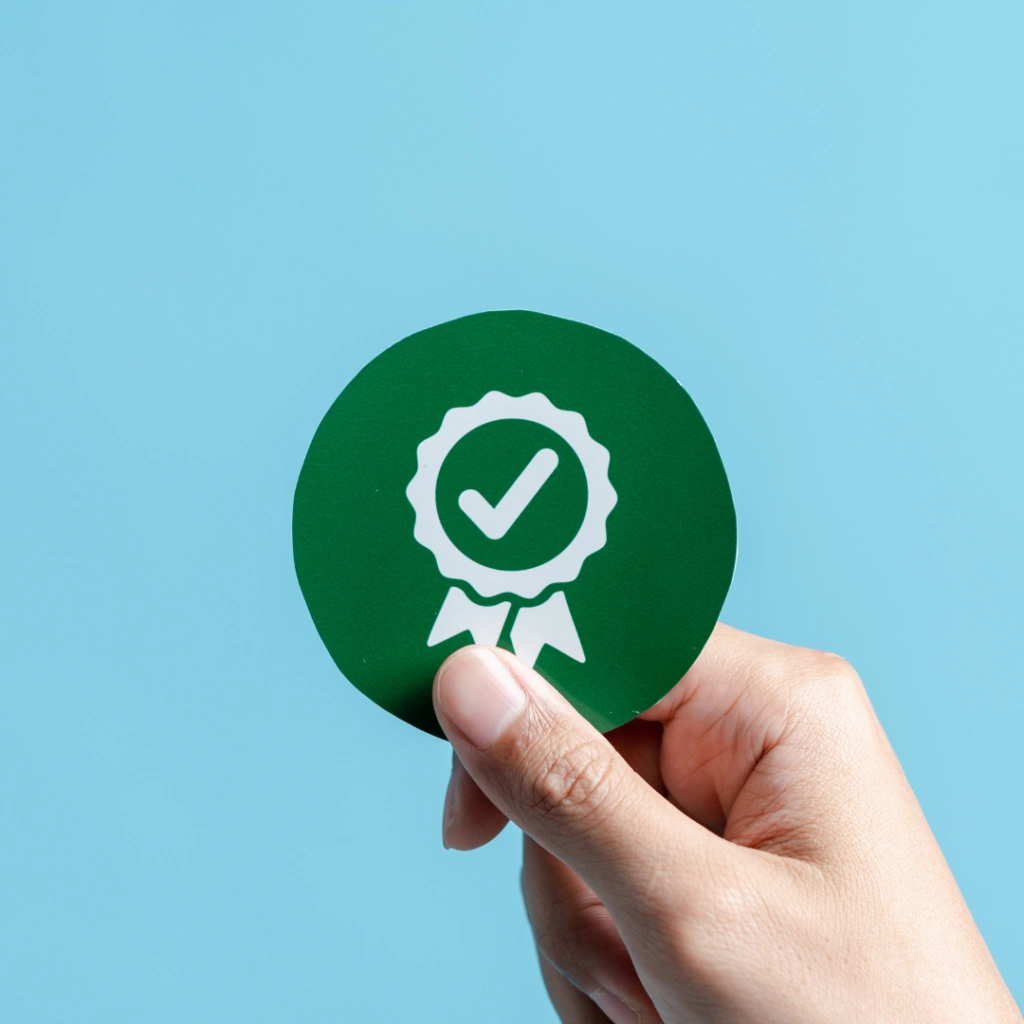 A picture of a hand holding a green paper circle with a medal graphic printed on. The graphic of the medal has a checkmark or tick inside it.