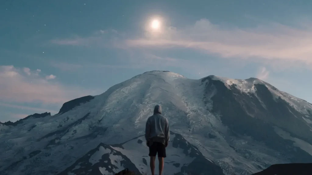 A man wearing a gray hoody stands looking at a snowy mountain in the distance as the sunsets