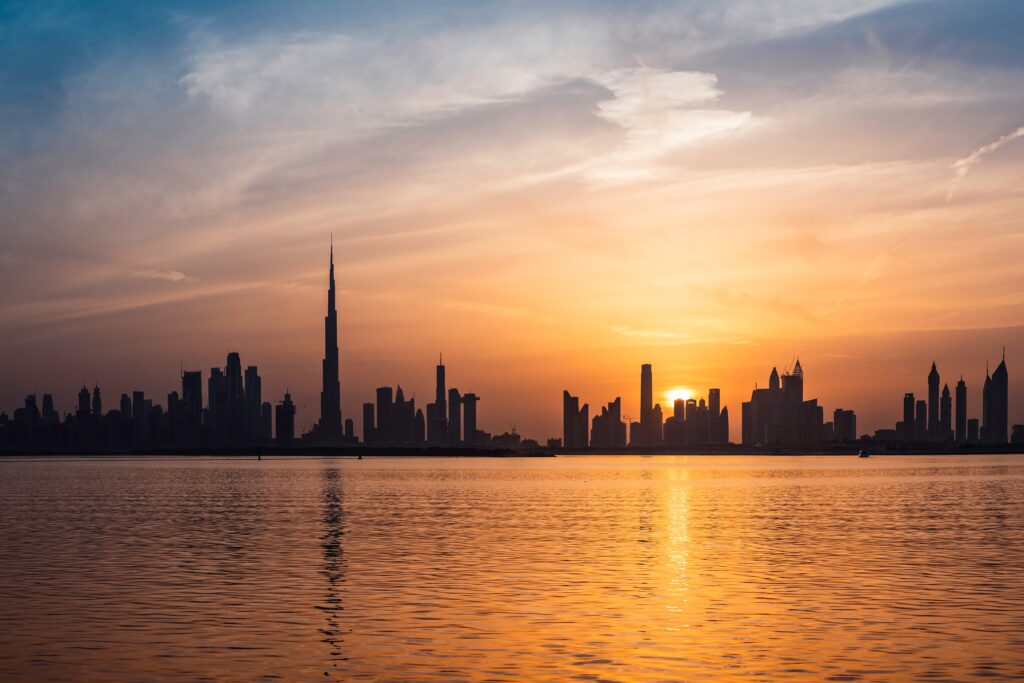 A stunning skyline of a city at sunset, featuring numerous high-rise buildings silhouetted against a vibrant orange and blue sky. The sun sets on the horizon, reflecting warmly on the calm water in the foreground.