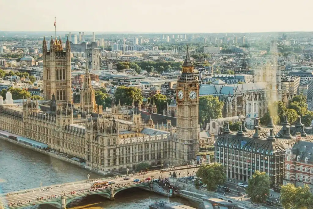 Aerial view of the Palace of Westminster and Big Ben in London on a sunny day. The River Thames and Westminster Bridge are in the foreground, while the cityscape stretches into the distance.