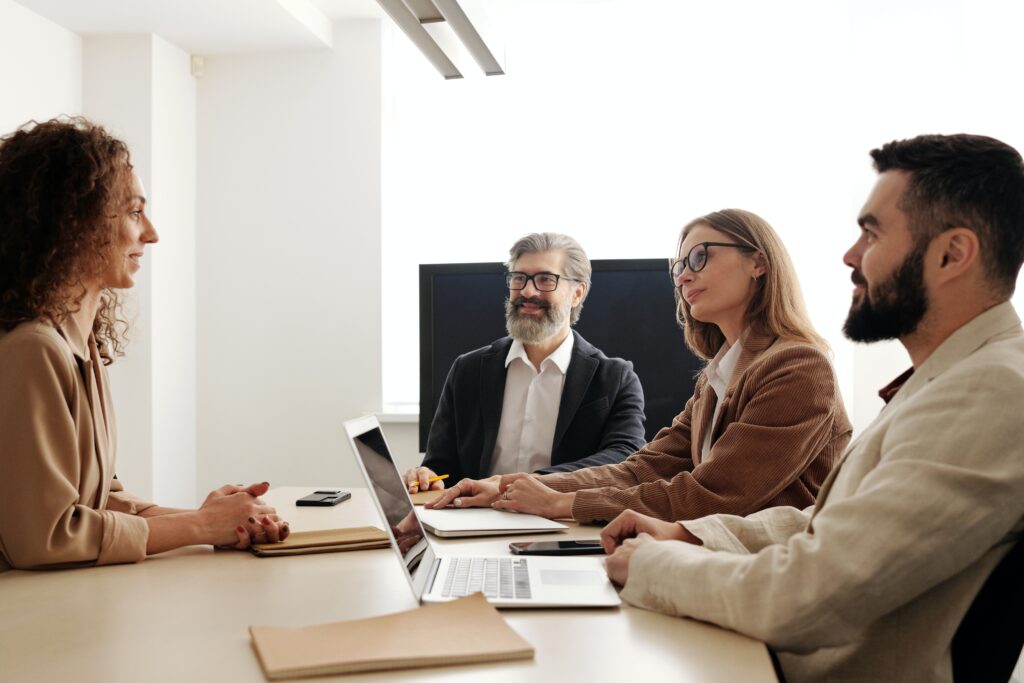 A bearded man with grey hair and glasses is smiling at a lady with long curly hair in a meeting room. Sitting across the table in the room are two other people on laptops.