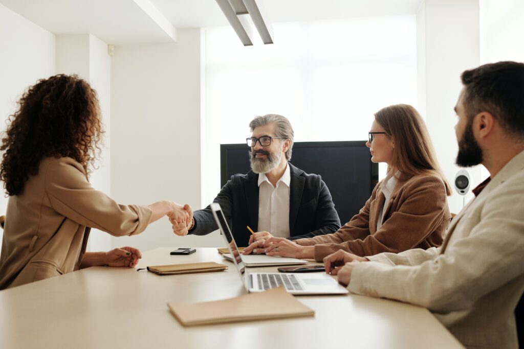 A bearded man with grey hair and glasses shakes the hand of a lady with long curly hair. They are sitting in a meeting room with two other people on a laptop.