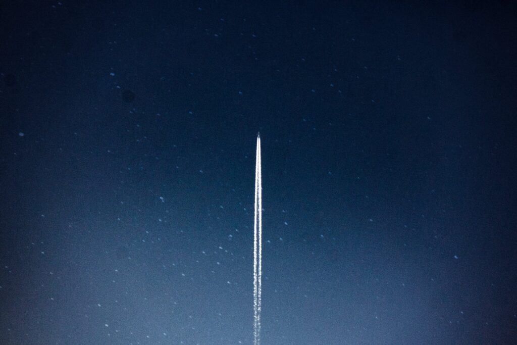 A view of the night sky, there's a multitude of stars in the background. In the centre a rocket can be seen flying upwards leaving a trail behind it.