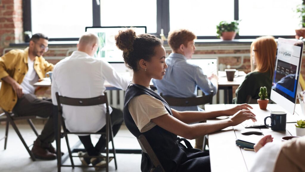 A diverse group of people working at desks with computers in a bright, modern office. One person is in the foreground typing, while others are engaged in various tasks. Large windows and green plants are visible, adding to the open atmosphere.