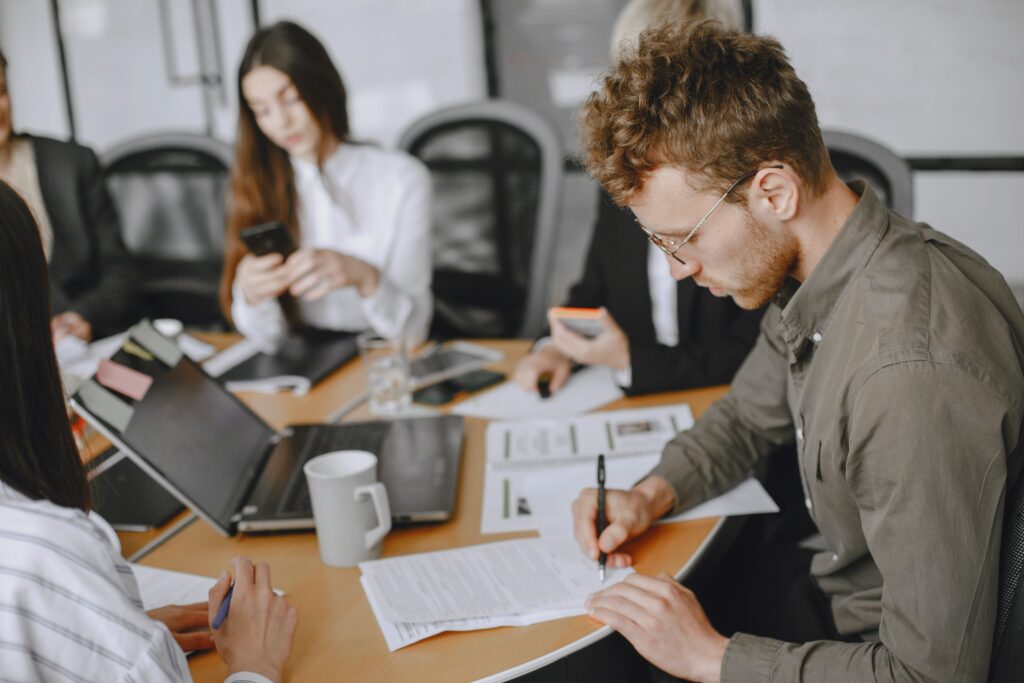 Four people are sitting around a conference table, engaged in work. One person is writing, while another checks their phone. Laptops, papers, and mugs are on the table, suggesting a business meeting or collaborative work session.