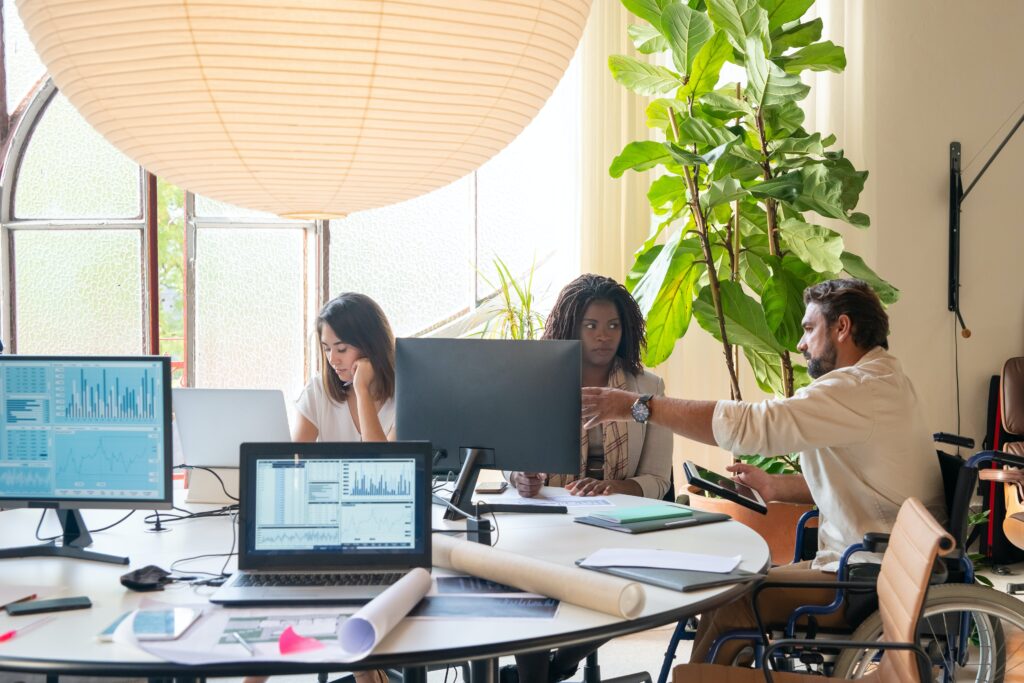 A diverse group of people work around a table with laptops and monitors. A man in a wheelchair points at a screen, while two women focus on their laptops. A large plant and soft lighting create a cozy atmosphere.