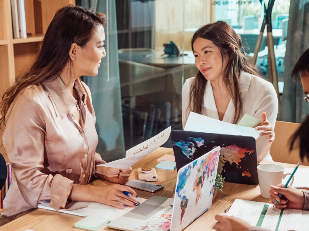 Two women are sitting at a table, engaged in a conversation. They are holding papers, and a laptop with a world map design is open in front of them. Coffee mugs and office supplies are scattered on the table, creating a collaborative workspace.