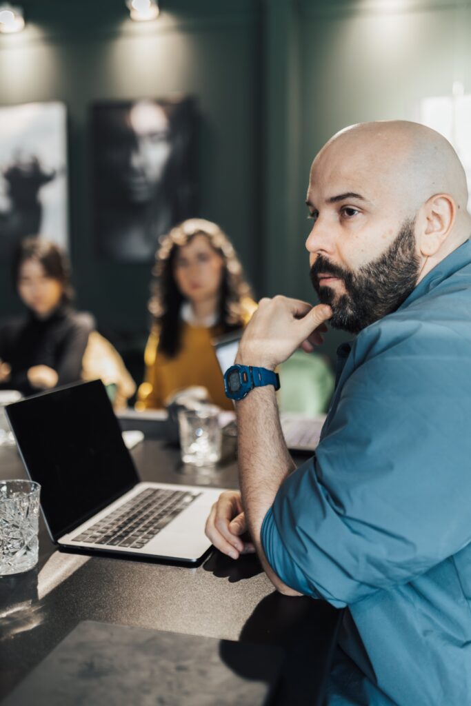 A bearded man seated at a table with a laptop open in front of him, resting his chin on his hand and looking thoughtful. Two blurred individuals are in the background, in a dimly lit room with artistic black-and-white portraits on the walls.