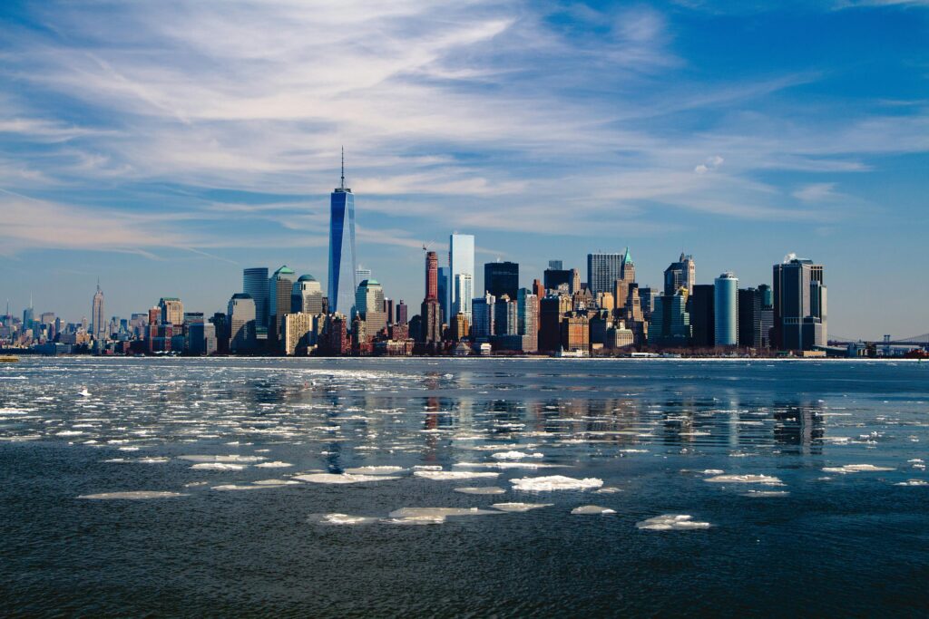 A clear view of the New York City skyline featuring tall skyscrapers, including One World Trade Center. The foreground shows icy waters with floating ice chunks, reflecting the cityscape. The sky is partly cloudy with bright sunlight.