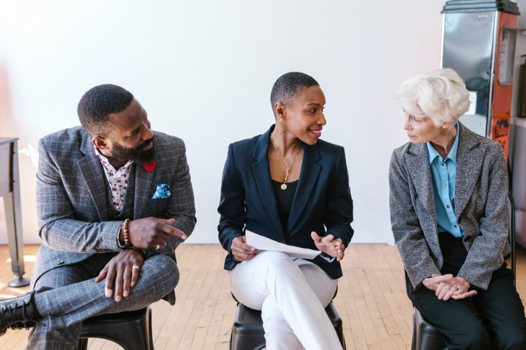 Three people in business attire sit on stools engaged in conversation. The person in the middle holds a paper. Theyre in a bright room with a wooden floor and white walls.