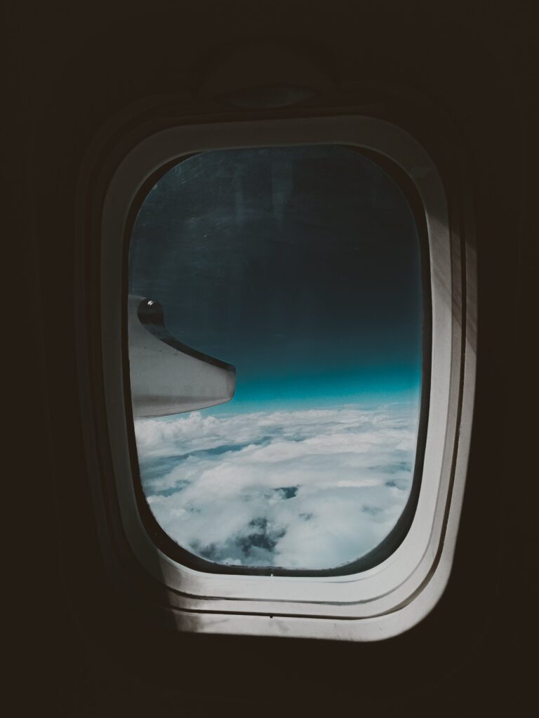 View through an airplane window, showing a wing against a backdrop of fluffy white clouds and a deep blue sky.