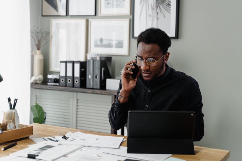 A man in a black shirt is sitting at a desk, talking on the phone and using a tablet. The desk is covered with papers. Behind him are a shelf with binders and a wall with framed artwork. The room has a modern, professional appearance.