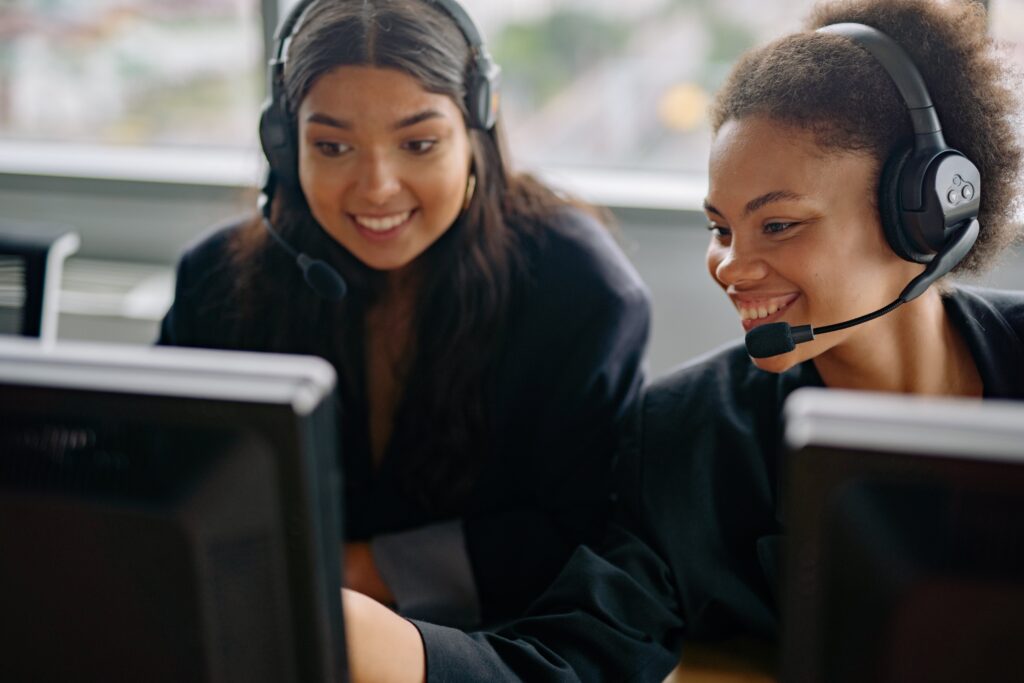 Two women wearing headphones that have mics and black business suits, smile while looking a computer monitor.