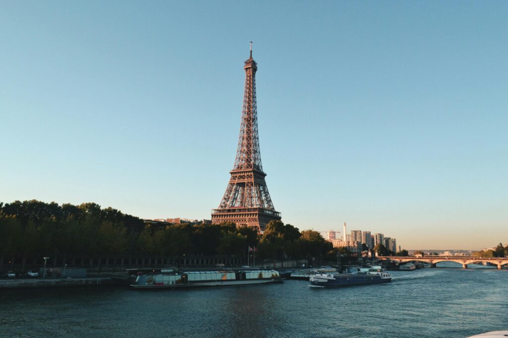 The Eiffel Tower in Paris, France, is captured during the day with a clear blue sky. The Seine River flows in the foreground with boats, and there are trees and buildings in the background.